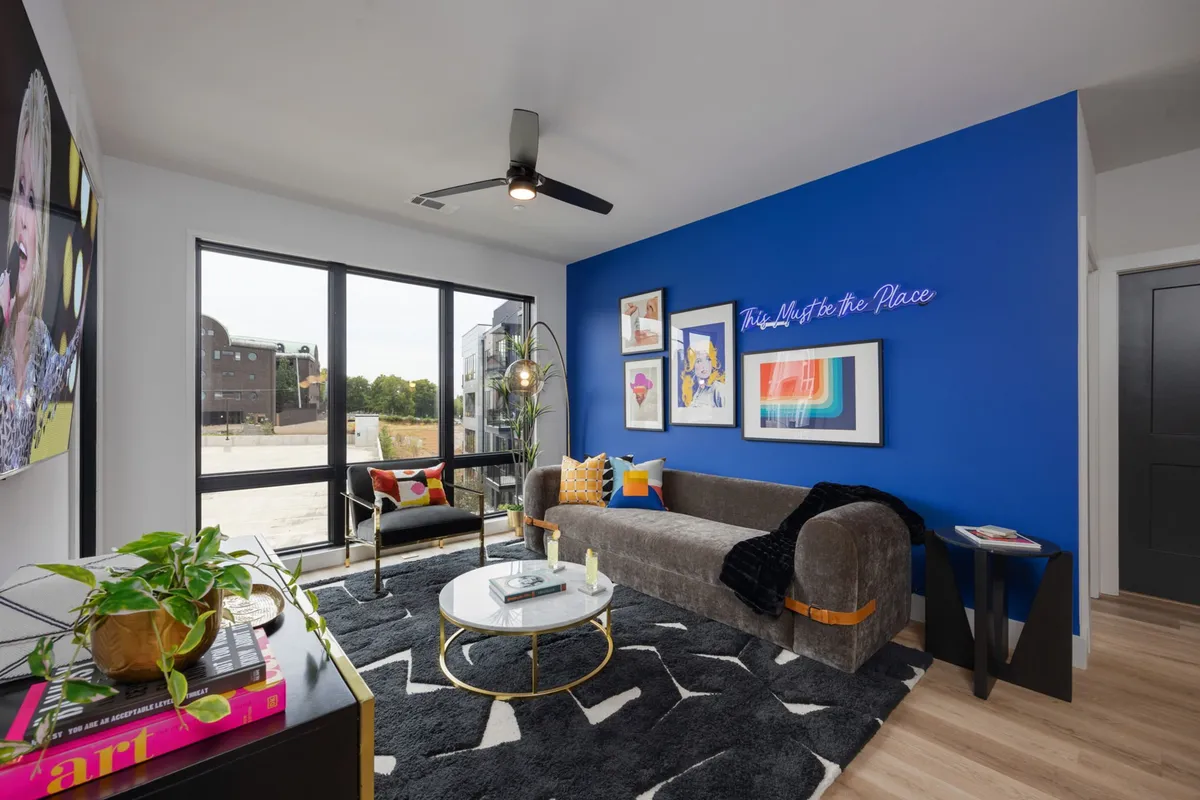 A cheerful living room featuring a blue accent wall, modern artwork, a stylish sofa, a decorative rug, and large windows that flood the space with natural light. The setup suggests a space for relaxation, entertaining, or enjoying a morning coffee while appreciating the view.
