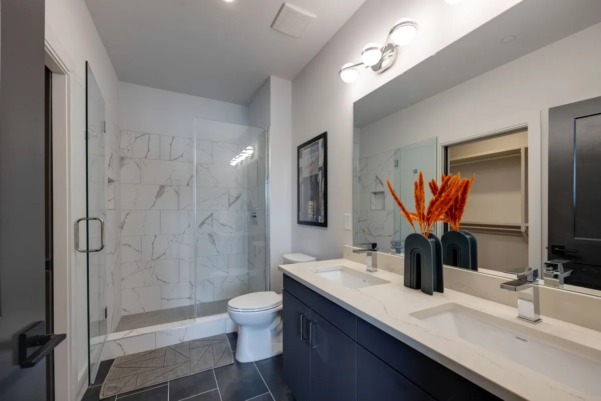 A fresh and modern bathroom featuring a double sink vanity, glass shower enclosure, and a decorative vase with dried flowers. The space is illuminated and inviting, evoking a spa-like ambiance for relaxation and rejuvenation.