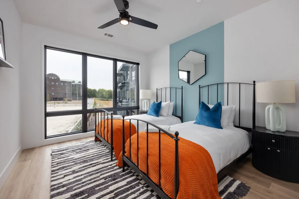 A bright, inviting bedroom with two twin beds adorned with orange throws, large windows letting in natural light, a decorative wall mirror, and modern bedside lamps. The room feels airy and perfect for families or friends to unwind.