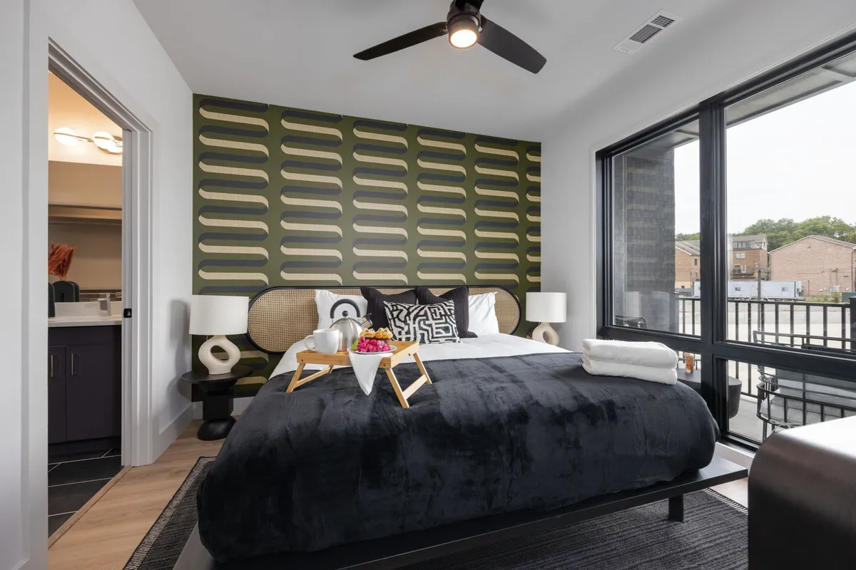 A spacious bedroom featuring a large bed with black bedding, bold patterned wallpaper, chic bedside lamps, and a decorative tray with fresh flowers. The atmosphere is contemporary yet cozy, with soft lighting and space for relaxation or reading.