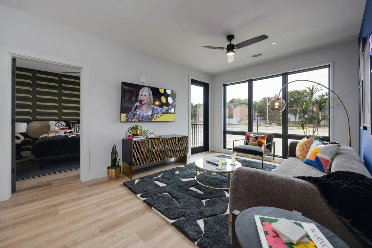 A bright, comfortable living area with a colorful couch, large windows bringing in abundant sunlight, a decorative rug, and a television. The atmosphere is lively and inviting, ideal for relaxing, watching shows, or hosting friends.