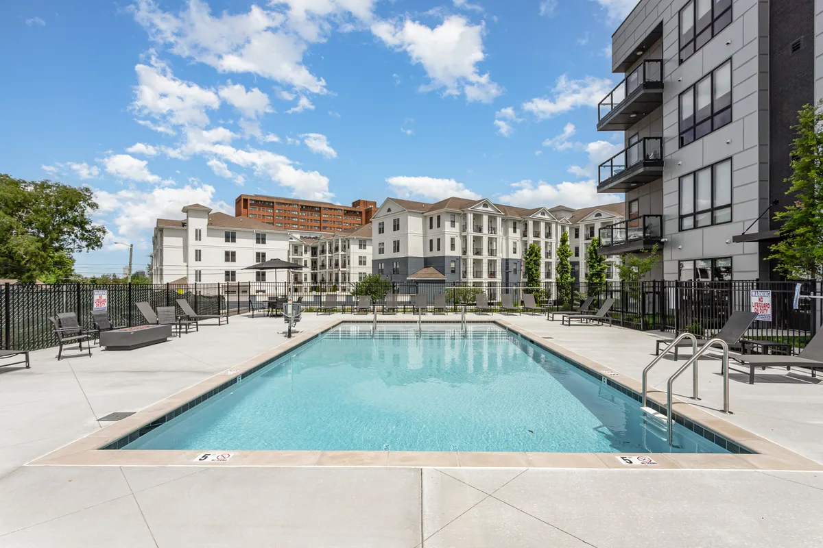 This exterior image captures a modern resort-style swimming pool surrounded by comfortable lounge chairs, lush landscaping, and sleek apartment buildings. The atmosphere is inviting and leisurely, suggesting a space perfect for relaxation, socializing, or sunbathing.