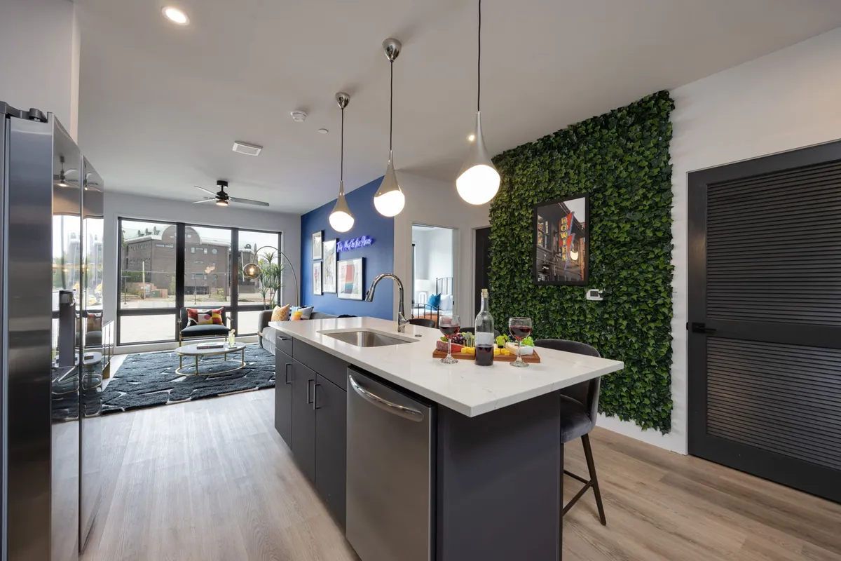 A light-filled kitchen scene with a spacious island, pendant lighting above, a touch of greenery from a living wall, and large windows that add to the airy, relaxed mood—suggesting a space perfect for socializing and group meals.